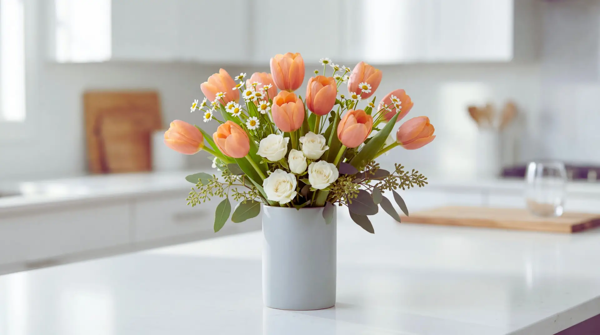 A coral tulip bouquet mixed with white spray roses, feverfew, and seeded eucalyptus in a pale gray vase on a bright kitchen island. Natural morning light and a softly blurred modern kitchen keep the scene clean and airy.