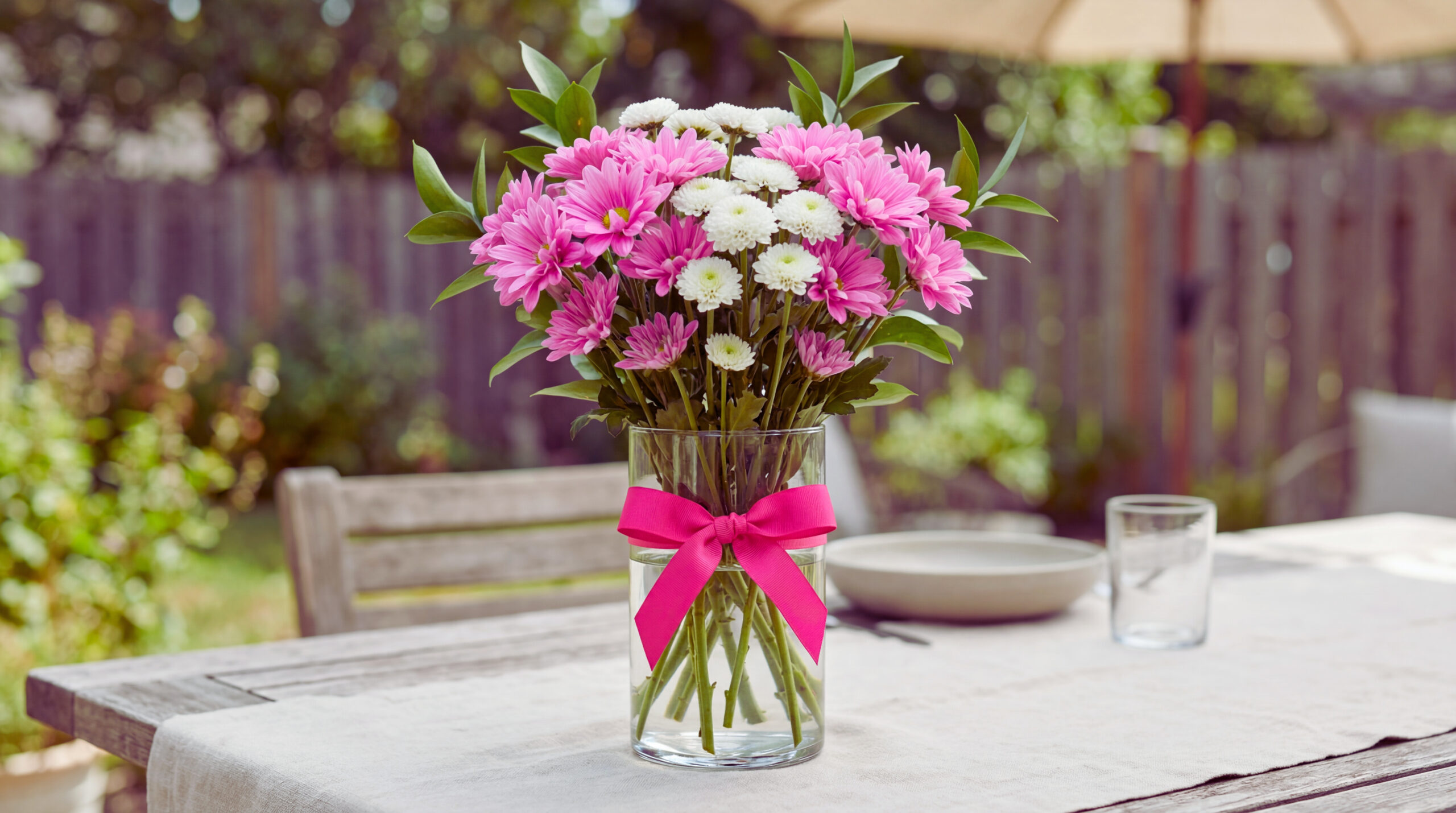 A bright pink chrysanthemum bouquet with white button mums in a clear vase on a patio table. The bouquet is loose and cheerful with minimal greenery in soft outdoor light.