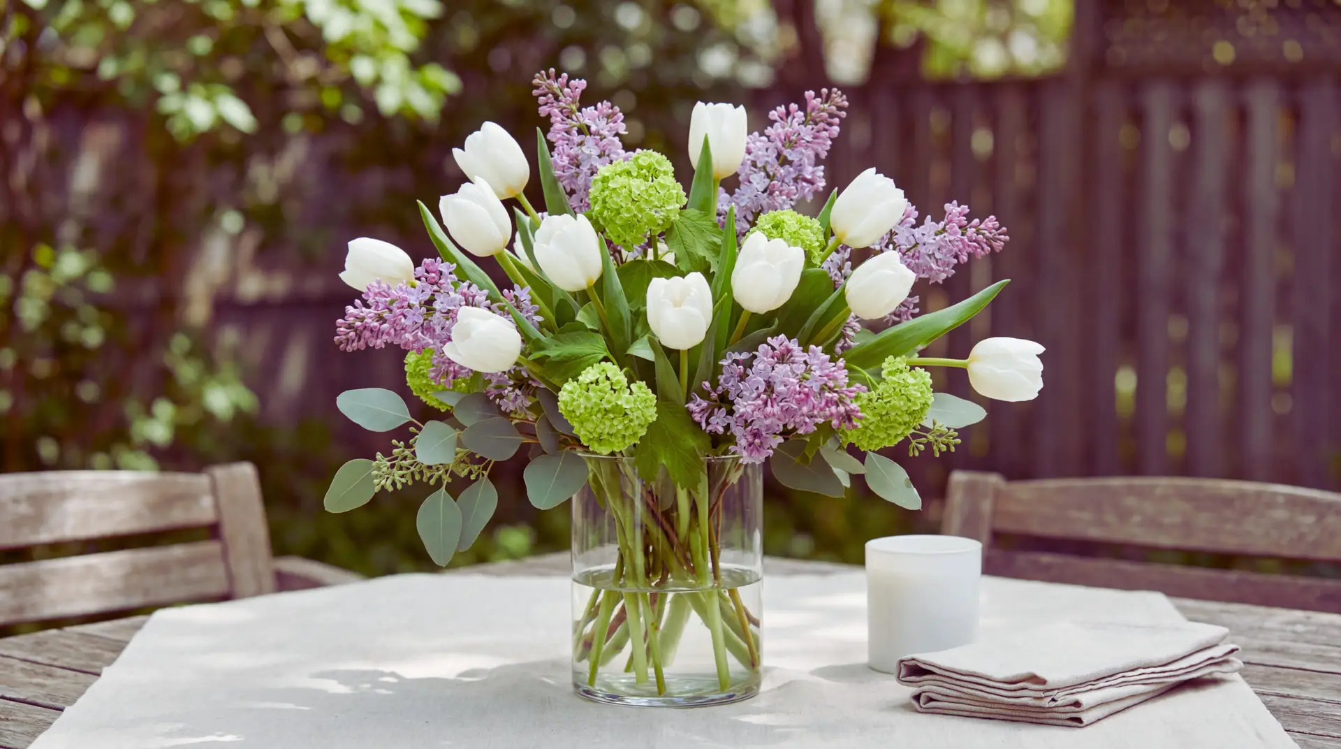 A white tulip bouquet mixed with lilac, viburnum, and seeded eucalyptus in a clear glass vase on a patio table. Soft outdoor shade and garden bokeh create a relaxed spring garden-party look.