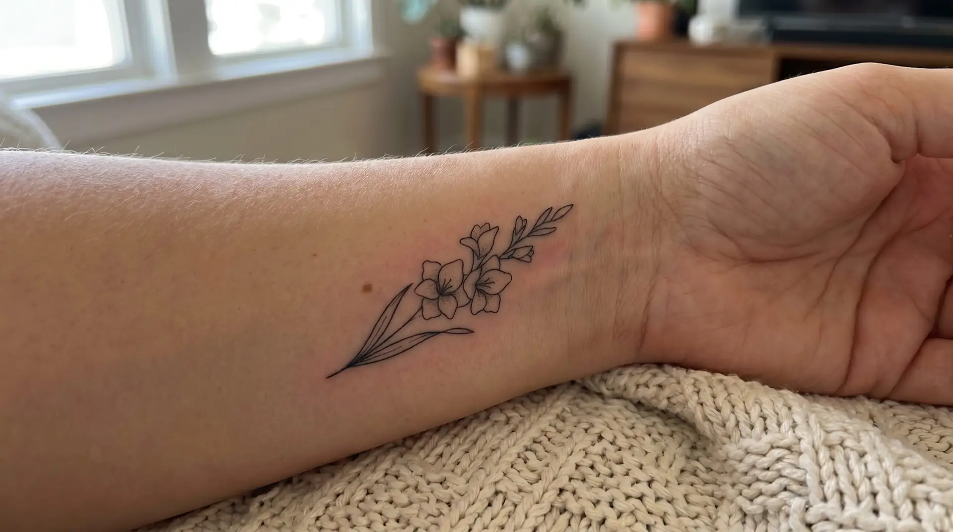 Small minimalist single-line gladiolus tattoo on inner wrist as August birth flower marker