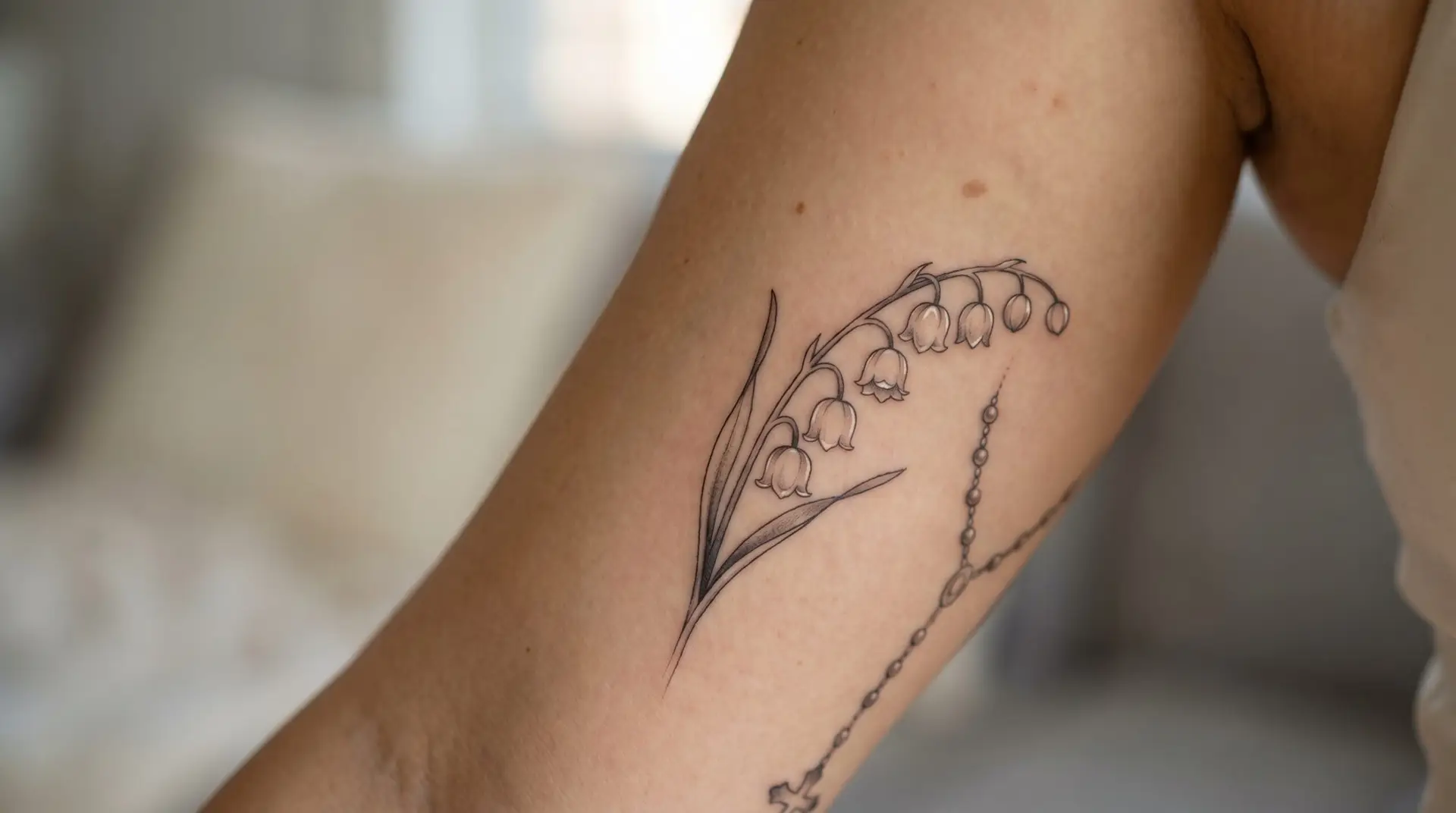 Marian commemorative tattoo on upper arm with lily of the valley stem and small rosary beads