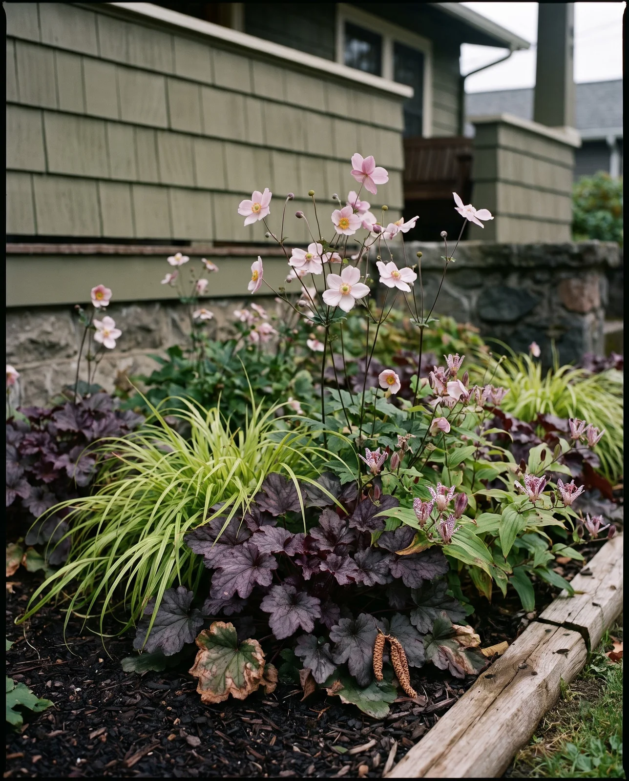 Shaded porch flower bed with pink Japanese anemones and heuchera.