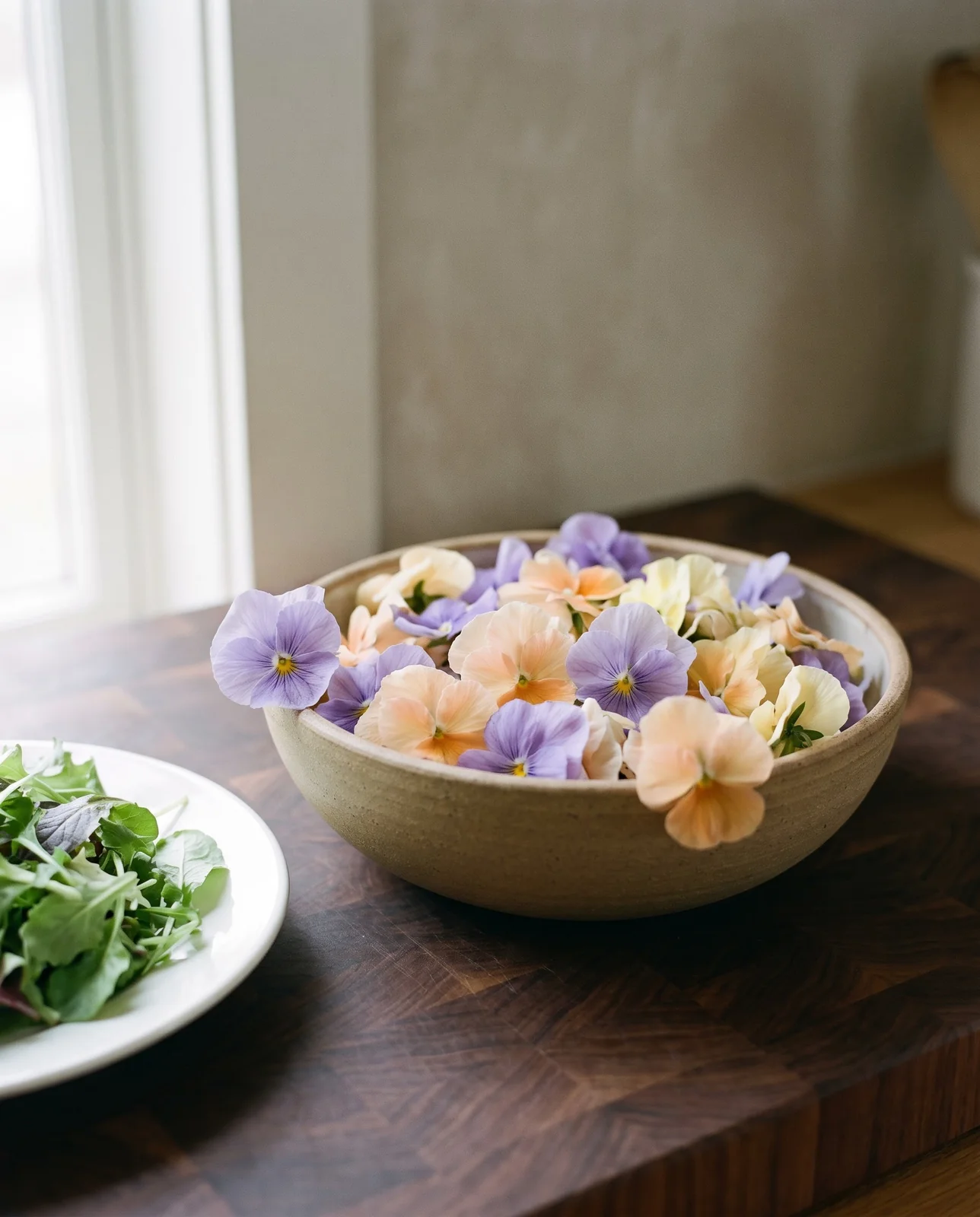Fresh edible pansy flowers in a ceramic bowl beside salad greens